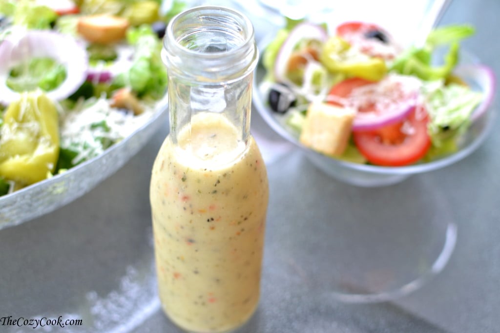 This homemade copycat Olive Garden Salad Dressing tastes just like the restaurant version. Learn how to make this salad at home for your family and friends! #copycatrecipe #restaurantcopycats #italianfood #italiansidedishes #sidedishideas #easyrecipes bottle of copycat olive garden salad dressing on a glass table with olive garden salad in the background.