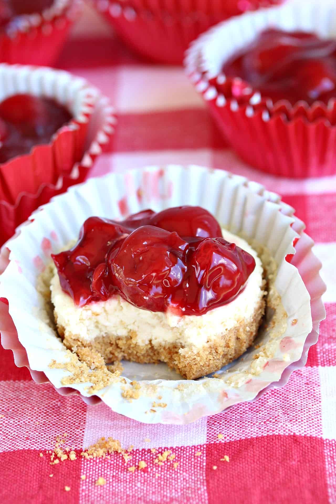 A sweet, buttery crust topped with creamy cheesecake and classic cherries. Ready in 35 minutes start to finish! A cherry cheesecake cupcake with a bite taken out of it on a red checkered surface.