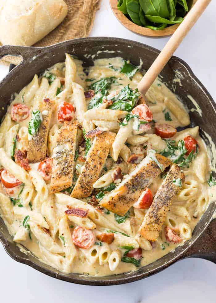 This loaded chicken bacon spinach pasta dish is tossed in a thick and creamy garlic Parmesan sauce. It's a perfect blend of flavors that will make you think you're eating at a restaurant! Chicken alfredo pasta in a black skillet with tomatoes and spinach.