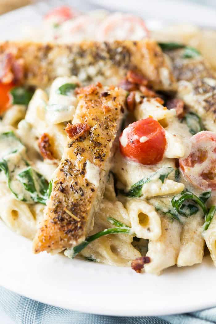 This loaded chicken bacon spinach pasta dish is tossed in a thick and creamy garlic Parmesan sauce. It's a perfect blend of flavors that will make you think you're eating at a restaurant! Close up image of seasoned and seared chicken next to a cherry tomato over pasta.