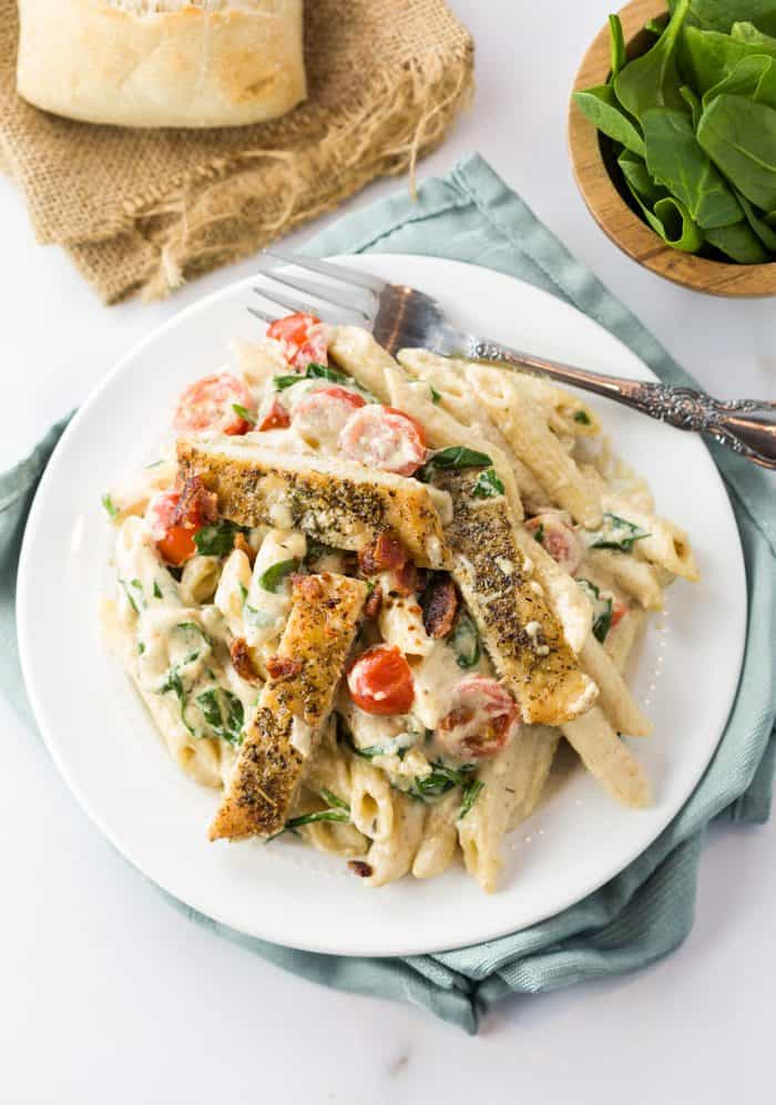 This loaded chicken bacon spinach pasta dish is tossed in a thick and creamy garlic Parmesan sauce. It's a perfect blend of flavors that will make you think you're eating at a restaurant! A white plate filled with chicken alfredo pasata on a table next to a roll.