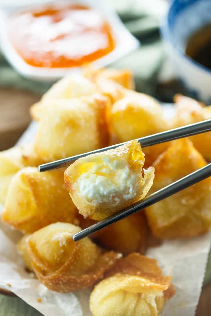 Crispy Wontons fried or baked to golden perfection and filled with a sweet, two-ingredient cream cheese filling. Chopsticks holding a fried wonton with cheese cheese filling inside and sweet and sour sauce in the background.