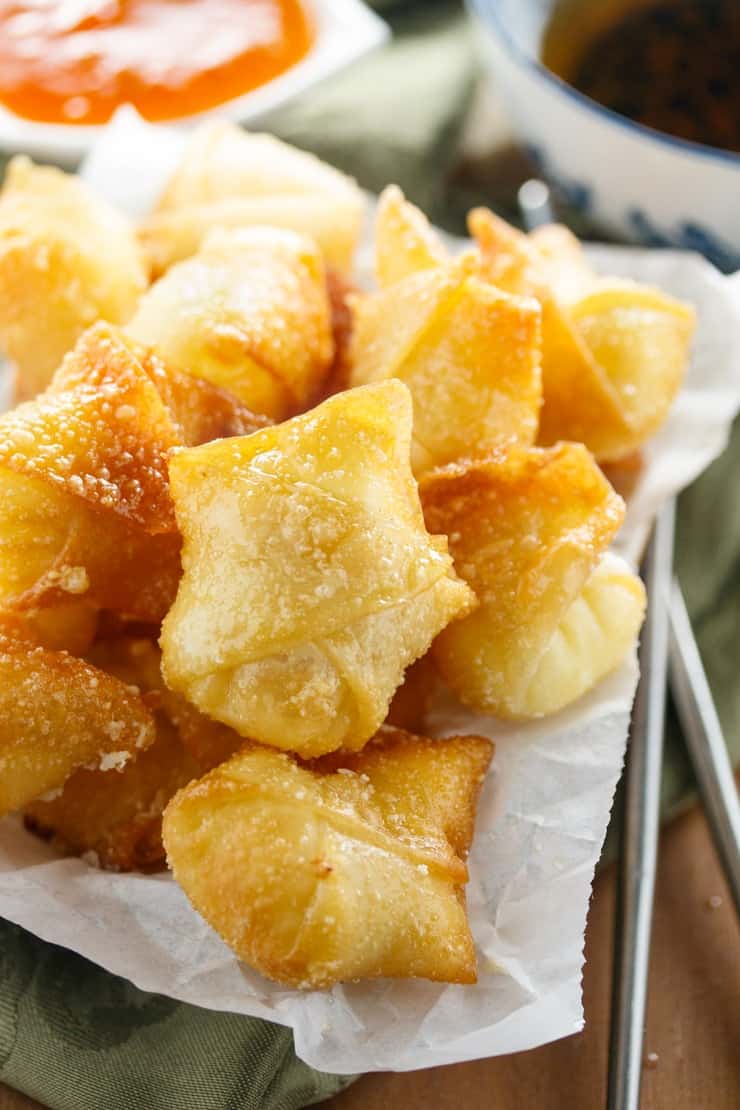 Crispy Wontons fried or baked to golden perfection and filled with a sweet, two-ingredient cream cheese filling. A pile of fried wontons with cream cheese filling and sweet and sour sauce in the background.