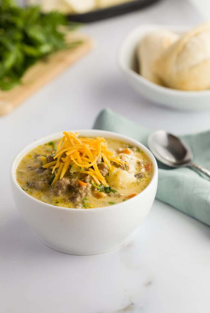 This slow cooker cheeseburger soup is loaded with tender ground beef and vegetables in a thick and creamy cheesy soup, the definition of comfort in a bowl! Slow Cooker Cheeseburger Soup in a white bowl on a white countertop with rolls in the background.