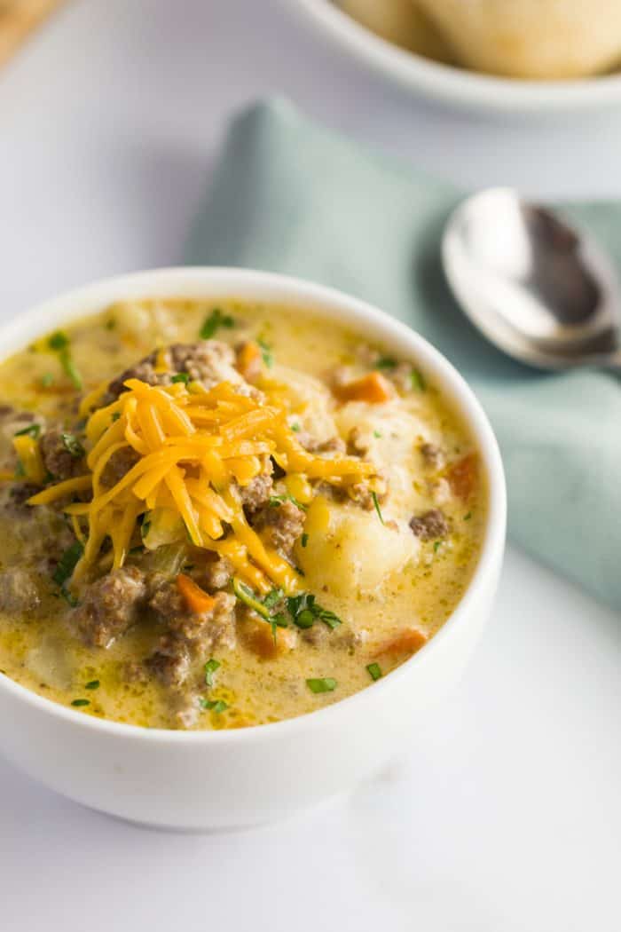This slow cooker cheeseburger soup is loaded with tender ground beef and vegetables in a thick and creamy cheesy soup, the definition of comfort in a bowl! Overhead view of Cheeseburger Soup topped with grated cheese on a white table.