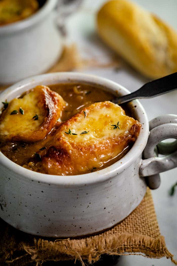 This traditional French Onion Soup has warm baguettes topped with hot, melted Gruyere cheese. High quality beef broth, a splash of wine, and perfectly caramelized onions are the secrets to this classic recipe. #easy #recipe #fallrecipes #winterrecipes #frenchcooking Two slices of baguette with melted cheese in a bowl of french onion soup and thyme on top.