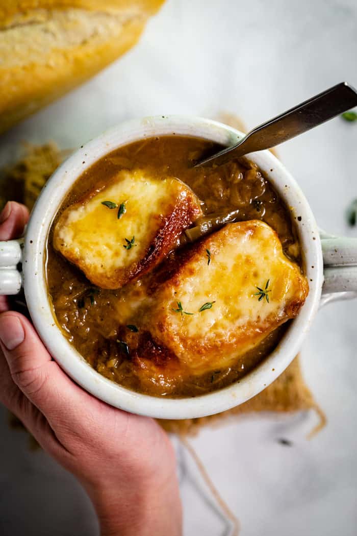 This traditional French Onion Soup has warm baguettes topped with hot, melted Gruyere cheese. High quality beef broth, a splash of wine, and perfectly caramelized onions are the secrets to this classic recipe. #easy #recipe #fallrecipes #winterrecipes #frenchcooking Overheat shot of a hand holding a white bowl filled with french onion soup topped with two cheesy baguettes.