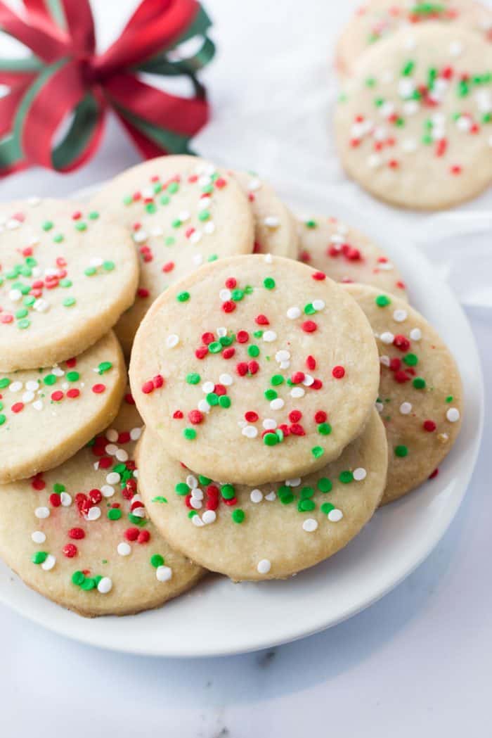 These soft, sweet, and buttery shortbread cookies only require 4 ingredients to make and are a classic holiday treat. Shortbread cookies on a white plate with a red holiday bow in background