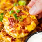 A hand holding a crispy slice of potato topped with melted cheese, bacon, and green onions.