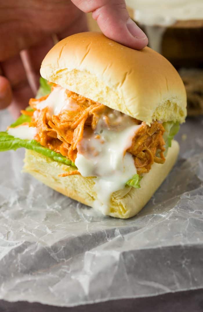 These slow cooker honey sriracha chicken sliders feature tender, slow cooked shredded chicken smothered in a sweet and tangy honey sriracha sauce, placed in a fresh slider roll and drizzled with savory blue cheese. | The Cozy Cook | #Sriracha #Chicken #Sliders #HoneySriracha #Sandwiches #SlowCooker #CrockPot #BlueCheese #BuffaloSauce A hand grabbing a slider bun filled with sriracha chicken drizzled with blue cheese.