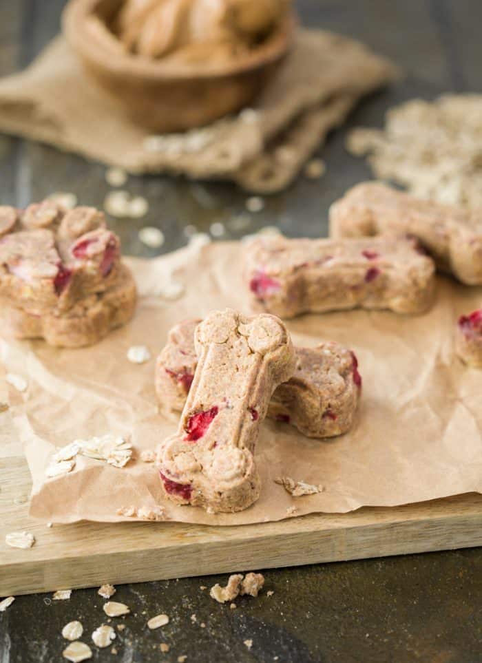 These homemade dog treats are loaded with strawberries, bananas, peanut butter, and oats. Everything you need to keep your dog happy and energized! homemade dog treats in the shape of a dog bone sitting on crinkled brown paper on a cutting board with oats.