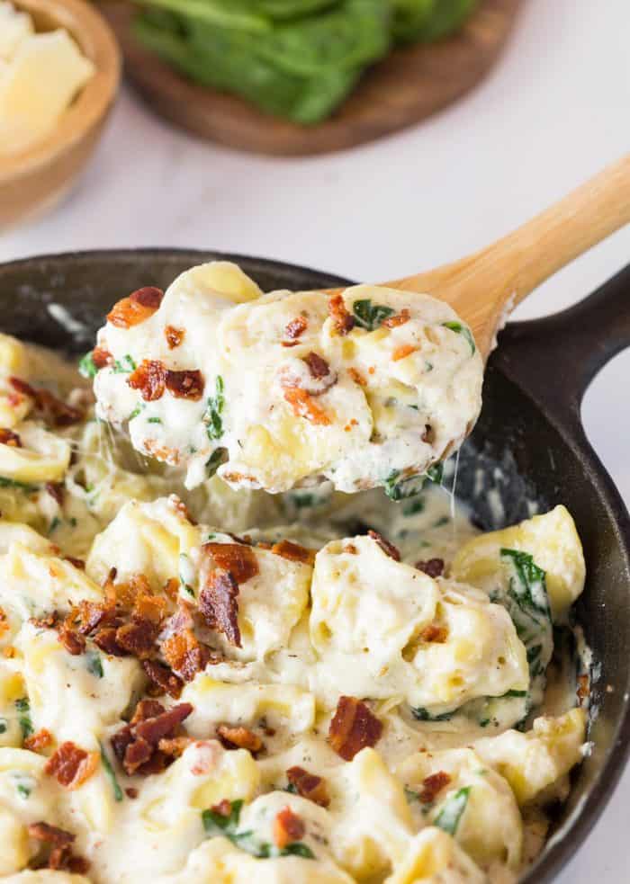This Tortellini Al Forno features stuffed tortellini tossed in a rich and creamy garlic cheese sauce and topped with crumbled bacon. | The Cozy Cook | #Tortellini #Pasta #OliveGarden #AlForno #Dinner, #Cheese #Parmesan #Bacon A cast iron skillet filled with tortellini in a cream sauce topped with bacon and a wooden spoon scooping some out.