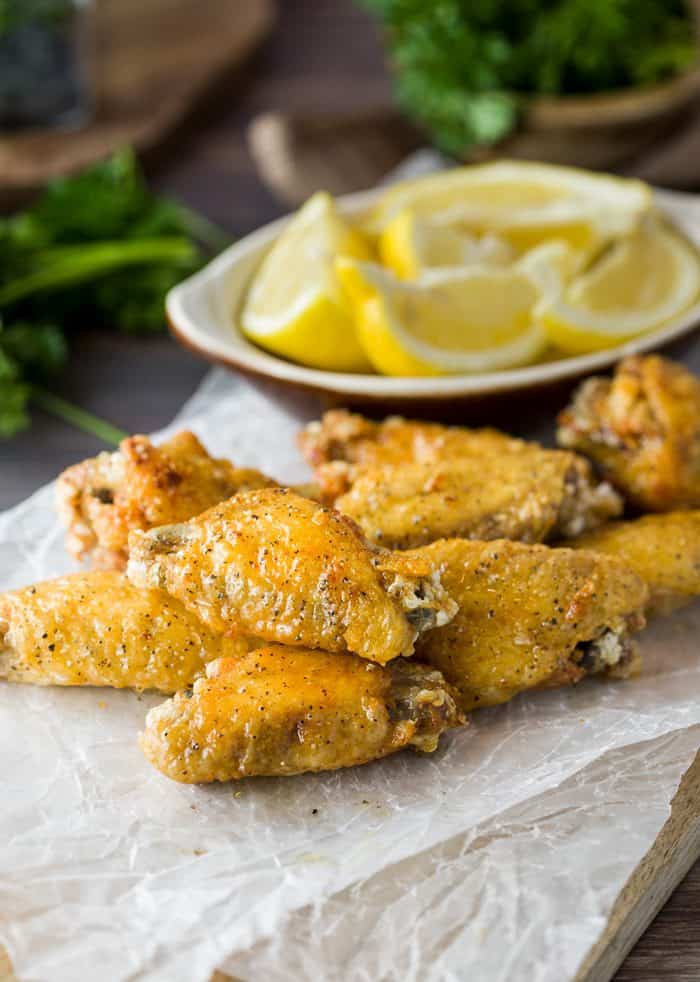 These lemon pepper wings are ultra crispy and brushed with a simple combination of lemon and butter, then topped with freshly ground pepper. Lemon Pepper Wings on a piece of white wax paper which a bowl of sliced lemons in the background.