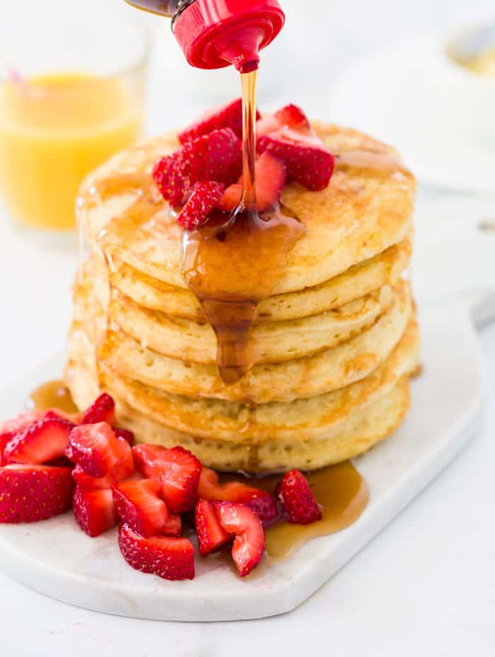 Sweet and fluffy pancakes served with fresh fruit and warm syrup. They taste just like IHOP's classic buttermilk pancakes, but are even better made at home! A tall stack of pancakes topped with strawberries with syrup being poured on top.