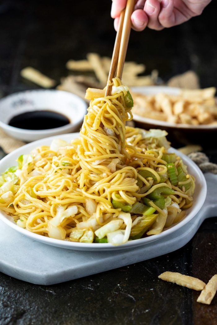This Copycat Panda Express Chow Mein tastes just like the restaurant version and is quick and easy to make right at home. In just 20 minutes you can enjoy this restaurant classic. Add seared shrimp, chicken, or beef to make it a protein-packed meal! | The Cozy Cook | #chowmein #copycat #pandaexpress #meatless #sidedish #chinesefood #restaurantcopycat #chinese #comfortfood #dinner #noodles A white plate on a marble cutting board filled with chow mein and a hand holding chopsticks twisting up the noodles.