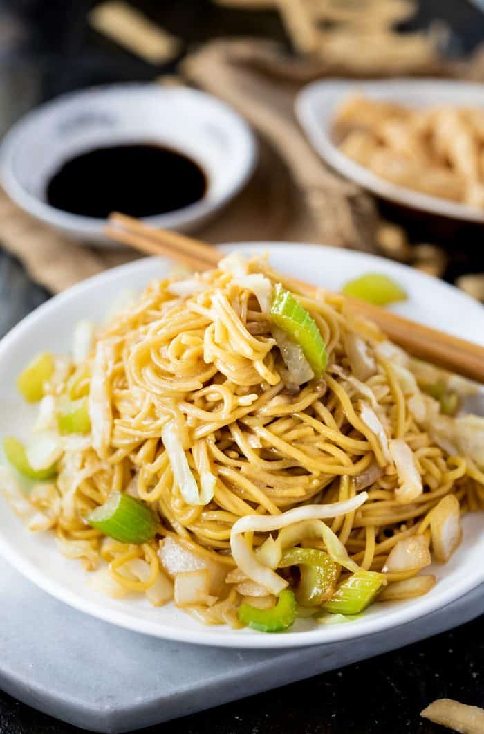 This Copycat Panda Express Chow Mein tastes just like the restaurant version and is quick and easy to make right at home. In just 20 minutes you can enjoy this restaurant classic. Add seared shrimp, chicken, or beef to make it a protein-packed meal! | The Cozy Cook | #chowmein #copycat #pandaexpress #meatless #sidedish #chinesefood #restaurantcopycat #chinese #comfortfood #dinner #noodles white plate filled with panda express chow mein with chopsticks and soy sauce in the background.