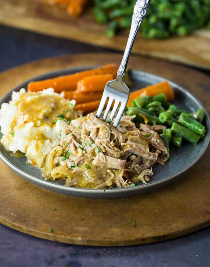 Just a few ingredients is all it takes to make this ultra-flavorful crock pot pork roast and gravy. The pork is perfectly seasoned and melts in your mouth when it’s time to eat! Shredded pork topped with parsley and gravy with a fork in it, with potatoes, carrots, and green beans in the background.