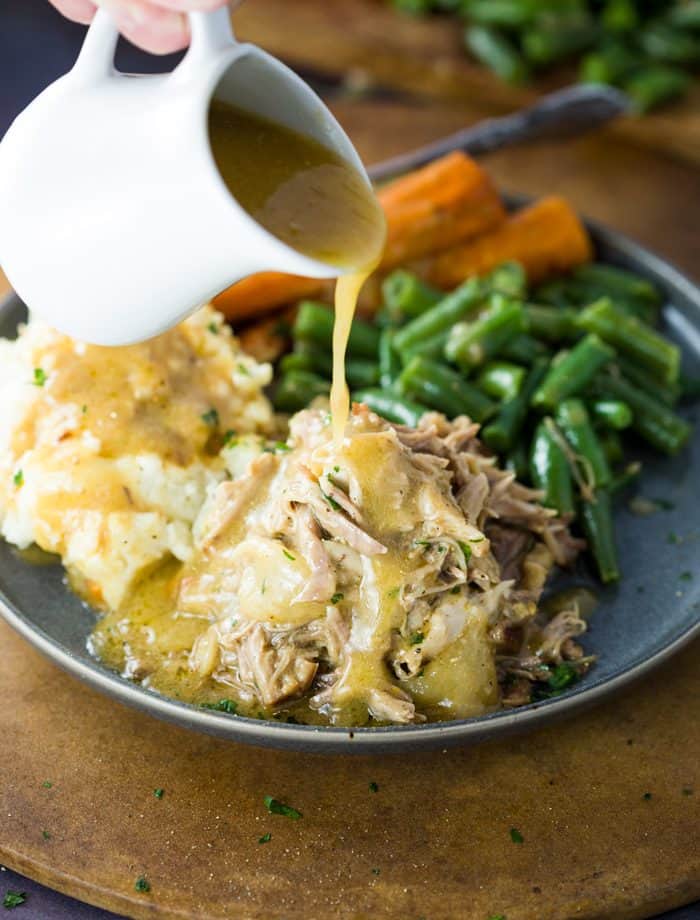 Just a few ingredients is all it takes to make this ultra flavorful crock pot pork roast and gravy. The pork melts in your mouth and is delicious with potatoes and vegetables. | The Cozy Cook | #pork #crockpot #slowcooker #dinner #comfortfood #meat #gravy #maindish #entree Gravy being poured over shredded pork on a plate with mashed potatoes, green beans, and carrots.