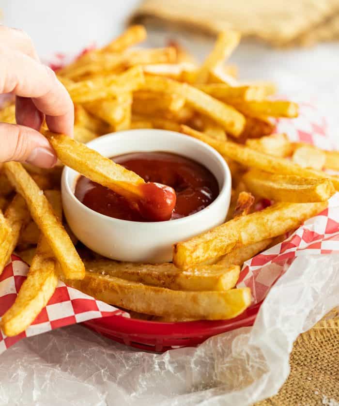 Learn the secret to making the best homemade french fries from russet potatoes! These are easy to make ahead of time and fry or bake in the oven for a quick snack or side dish idea! Hand dipping crispy homemade french fry into a white bowl of ketchup inside a basket of fries