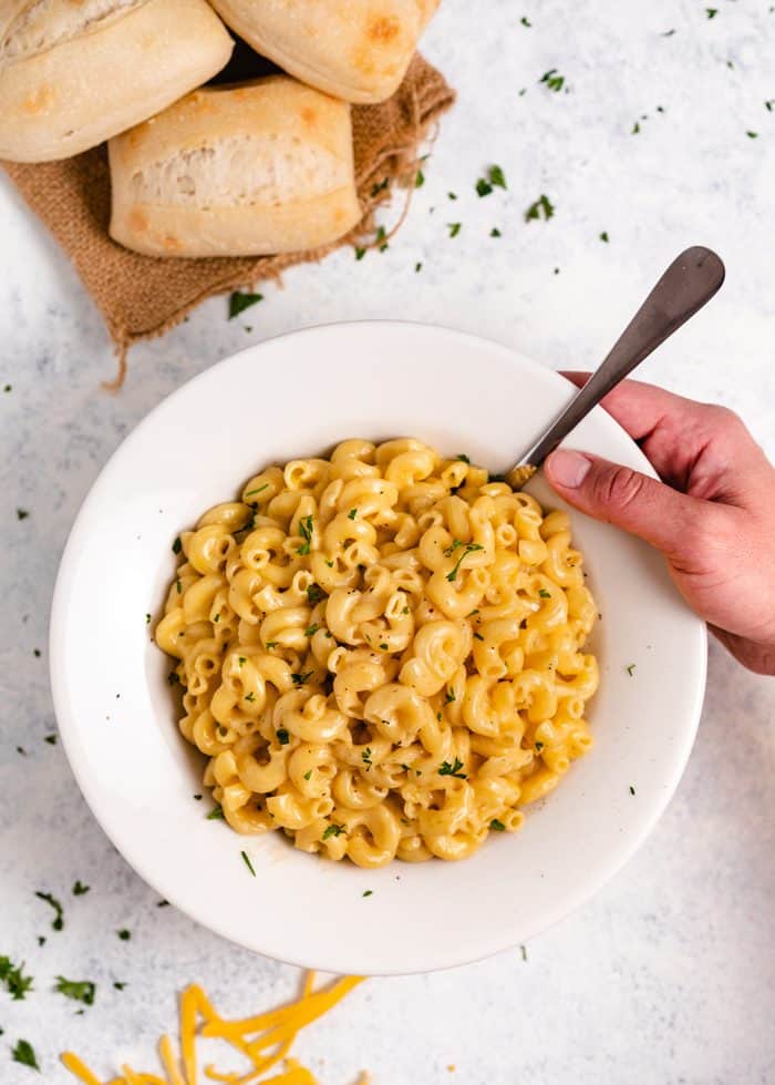 This super creamy Instant Pot macaroni and cheese cooks in just 5 minutes with only a single pot to clean! Your whole family will love this extra easy, cheesy meal! | The Cozy Cook | #macaroni #cheese #instantpot #dinner #pasta A hand reaching for a white bowl filled with macaroni and cheese on a light white surface with rolls in the background.