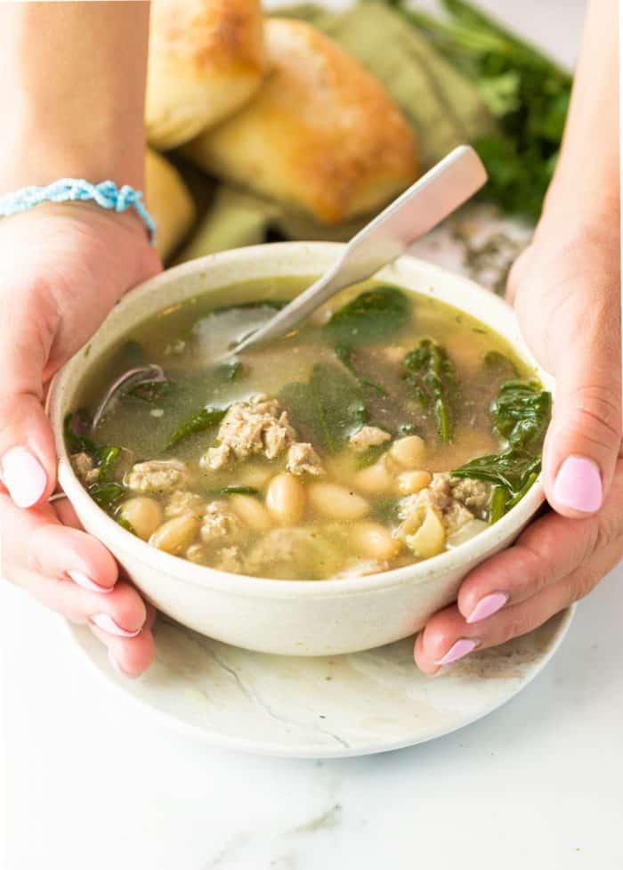 Ground Turkey simmered in savory chicken broth with white beans, spinach, and simple seasonings. A healthy, protein-packed meal that will leave you feeling comforted and satisfied! | The Cozy Cook | #soup #turkey #healthy #broth #whitebeans #protein #fall Two hands holding a white bowl filled with warm soup with ground turkey, white beans, and spinach in broth.