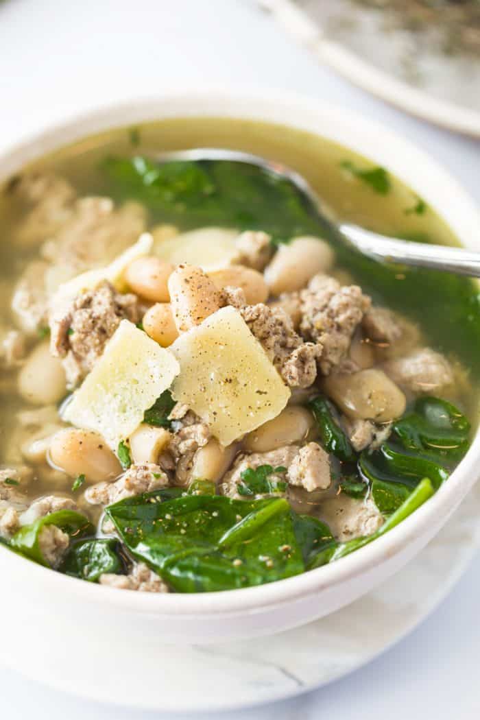 Ground Turkey simmered in savory chicken broth with white beans, spinach, and simple seasonings. A healthy, protein-packed meal that will leave you feeling comforted and satisfied! | The Cozy Cook | #soup #turkey #healthy #broth #whitebeans #protein #fall Ground Turkey, Spinach, and White Bean Soup in white bowl with Parmesan flakes on top with ground pepper.