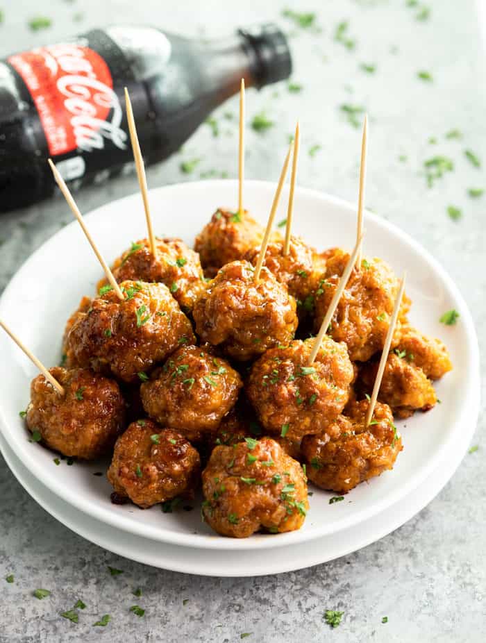 These spicy sausage balls come together with nothing more than cheddar, cream cheese, sausage, and bisquick! Make them ahead of time and keep them warm in the Crock Pot! #christmasappetizers #appetizerideas #fingerfood #newyearseverrecipes A white plate filled with brown glossy spicy sausage bites with a coca cola bottle in the background.