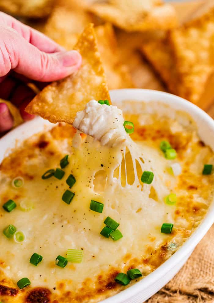 a fried wonton chip being dipped into a white bowl of crab rangoon dip, pulling up the warm cheese.
