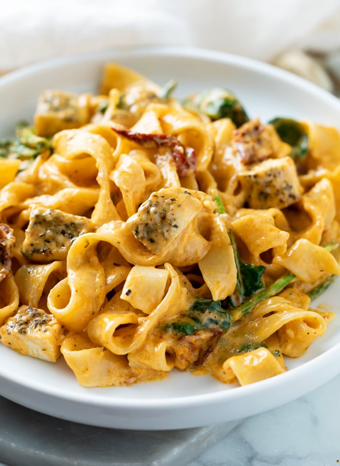 A white plate with Creamy Tuscan Chicken Pasta with noodles, chicken, sundried tomatoes, and spinach.