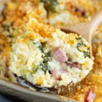 A wooden spoon scooping up ham and rice casserole with cheese from a casserole dish.