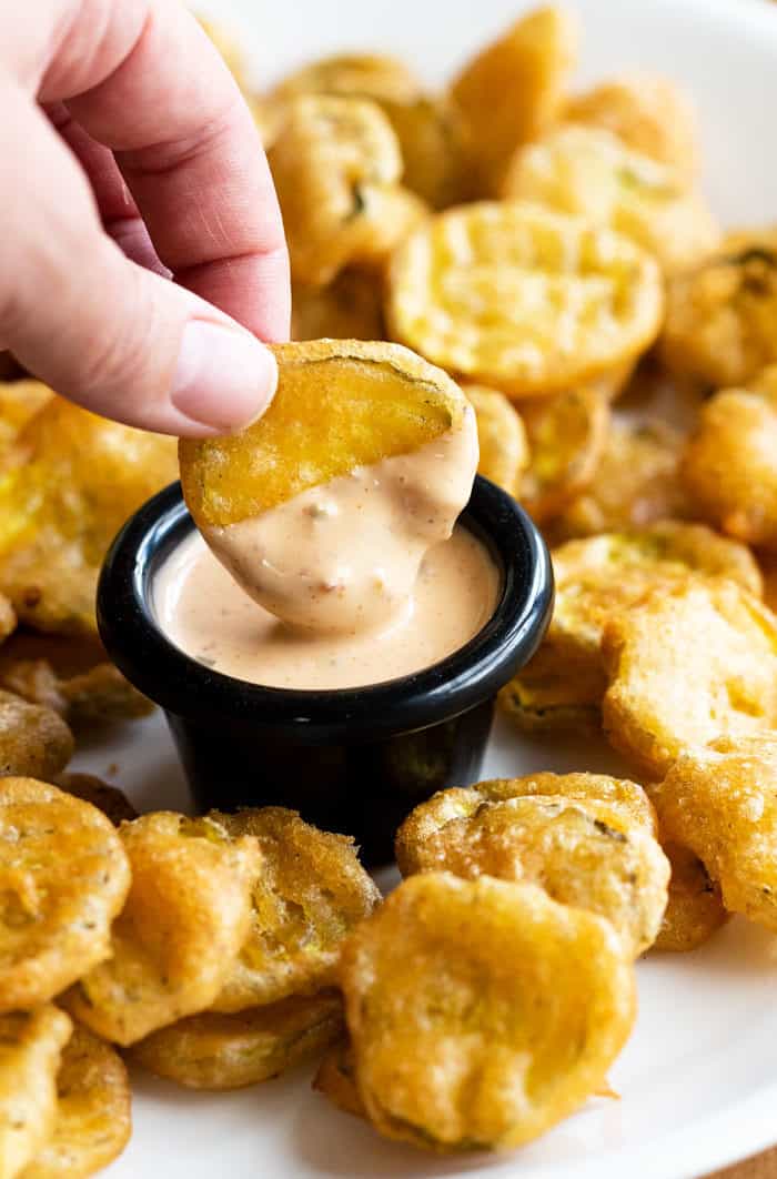 A hand dipping a golden fried pickle into a black ramekin of dipping sauce.