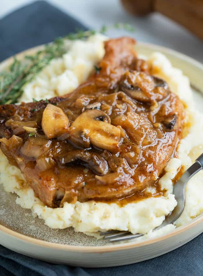 Seasoned Pork Chops are smothered in a thick and flavorful French Onion sauce with gravy and mushrooms. They are pan fried or baked in the oven. Top with hot, melted cheese for extra indulgence! #smotheredporkchops #onepotmeals #mushroomsauce #caramlizedonions #pork White potatoes on a plate topped with a pork chop smothered in a french onion sauce with mushrooms.
