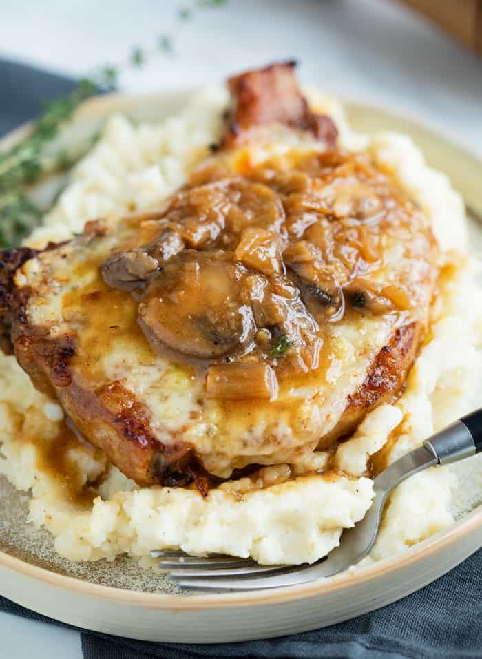 Seasoned Pork Chops are smothered in a thick and flavorful French Onion sauce with gravy and mushrooms. They are pan fried or baked in the oven. Top with hot, melted cheese for extra indulgence! #smotheredporkchops #onepotmeals #mushroomsauce #caramlizedonions #pork A Pork Chop on top of mashed potatoes topped with melted cheese and a french onion mushroom sauce.