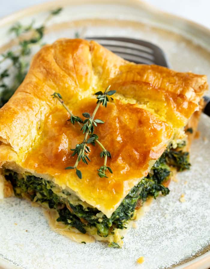 This spinach casserole recipe is made with eggs, caramelized onions, gruyere cheese, and golden puff pastry. An easy side dish idea for Thanksgiving or a family dinner! #makeahead #creamy #sidedishes #holidaysidedish #christmassidedish #vegetablesidedishes #comfortfood A slice of spinach pie with a golden brown puff pastry topping garnished with fresh thyme on a plate.