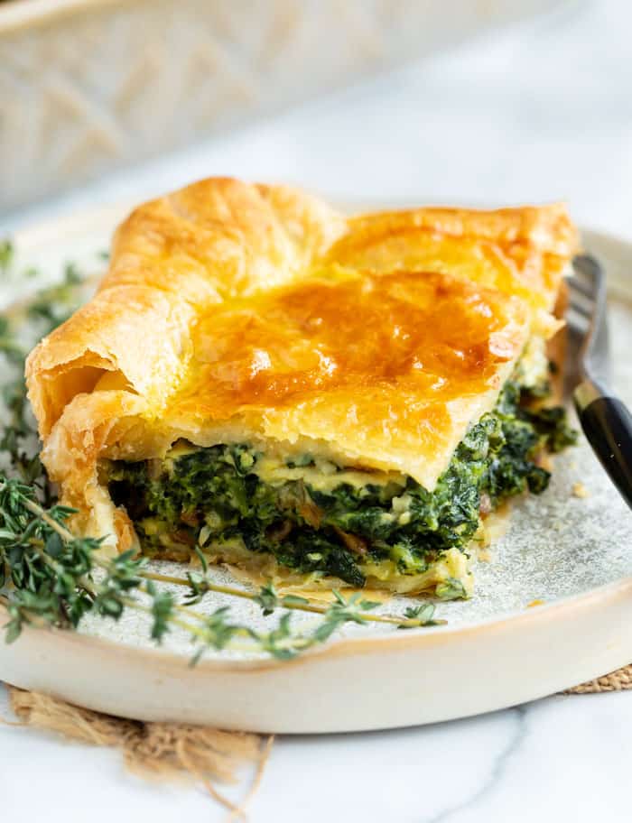 This spinach casserole recipe is made with eggs, caramelized onions, gruyere cheese, and golden puff pastry. An easy side dish idea for Thanksgiving or a family dinner! #makeahead #creamy #sidedishes #holidaysidedish #christmassidedish #vegetablesidedishes #comfortfood A slice of spinach pie on a plate with fresh thyme next to it and a fork on the side.