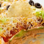 A tortilla chip on top of 7 layer dip in a baking dish.