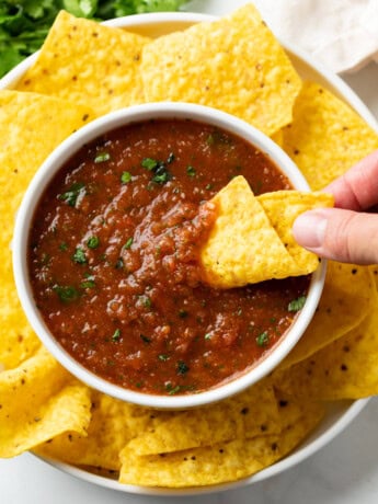 A hand dipping a tortilla chip into a white bowl of salsa with tortilla chips around it.
