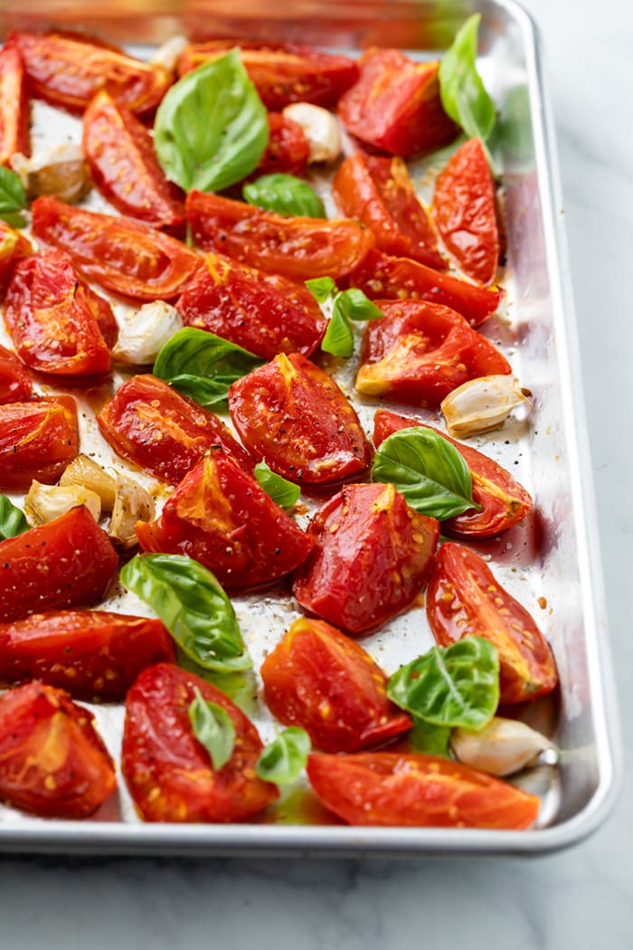 A rimmed baking sheet with roasted tomatoes and basil on top with salt and freshly cracked pepper.