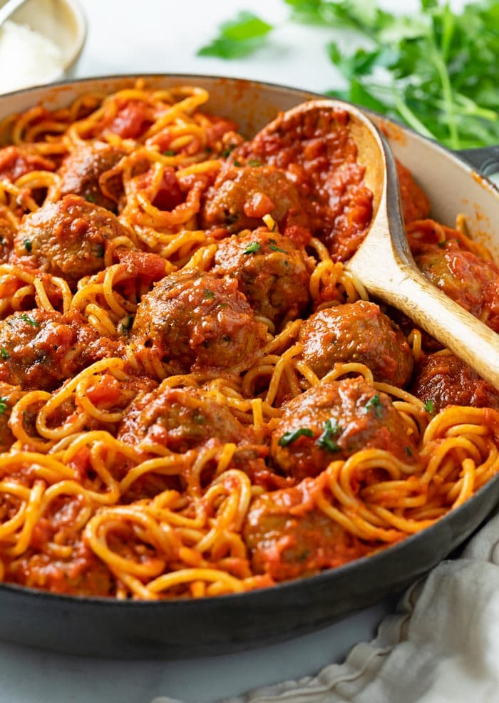 A pot of Spaghetti and Meatballs with a wooden spoon on the side and parsley in the background.