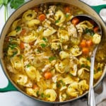 A soup pot filled with Chicken Tortellini Soup with vegetables, seasoning, and fresh parsley.