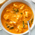 A bowl of creamy Tomato Tortellini Soup with spinach in it and a spoon on the side.
