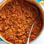 A Dutch oven filled with Meaty Chili with Kidney beans, peppers, and a chili sauce with a ladle on the side.