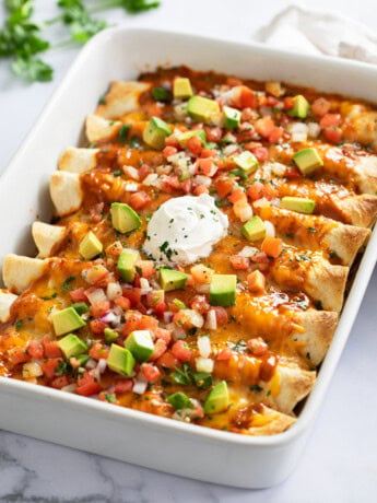 Beef Enchiladas in a white baking dish topped with cheese, tomatoes, avocados, and sour cream.