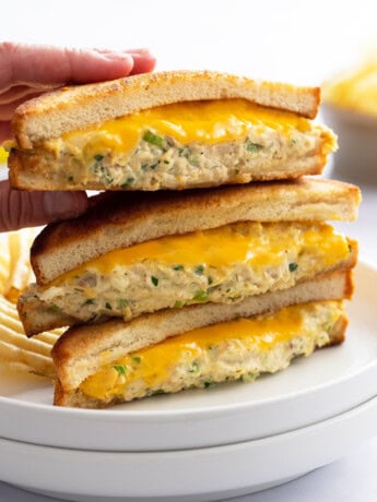 A hand taking the top Tuna Melt from a stack of 3 on a white plate with melted cheese.