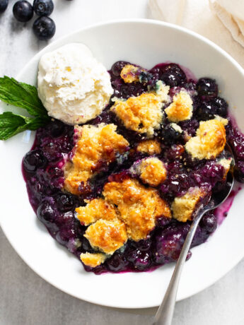 Blueberry Cobbler in a white bowl with a scoop of vanilla ice cream and a sprig of mint on the side.
