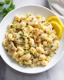 Chicken Piccata Pasta in a white bowl with chopped parsley on top and sliced lemon on the side.