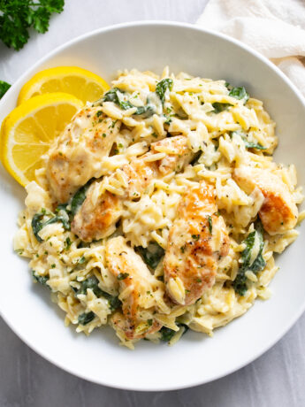 Lemon Chicken Orzo in a white bowl with lemon slices on the side and chicken and spinach in a creamy sauce with orzo.
