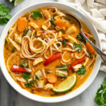 Thai Chicken Soup with noodles, chicken, and vegetables in an orange broth with garnishes.