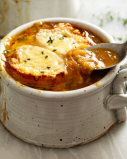 A crock of French Onion Soup with melted cheese and bread on top and a spoon scooping up some of the rich broth on the side.